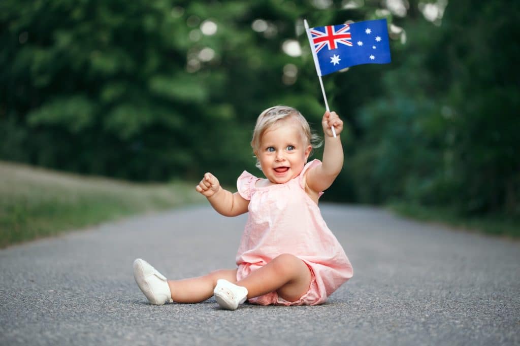 Cute baby girl sitting on ground road street outdoor waving Australian flag. Australia Day holiday.