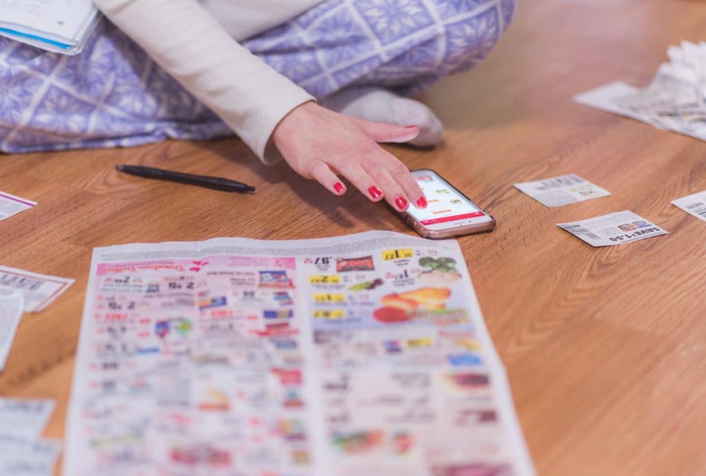 Millennial woman looking for coupons in newspapers with aid of smartphone